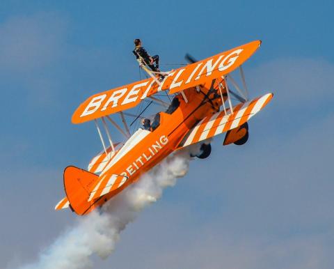 Breitling orange biplane ascending in blue sky trailing smoke with stunt performer standing on wing.